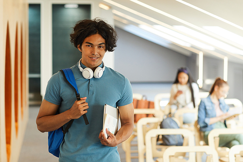 Portrait of smiling handsome student guy with messy hair standing with satchel and laptop in modern university room