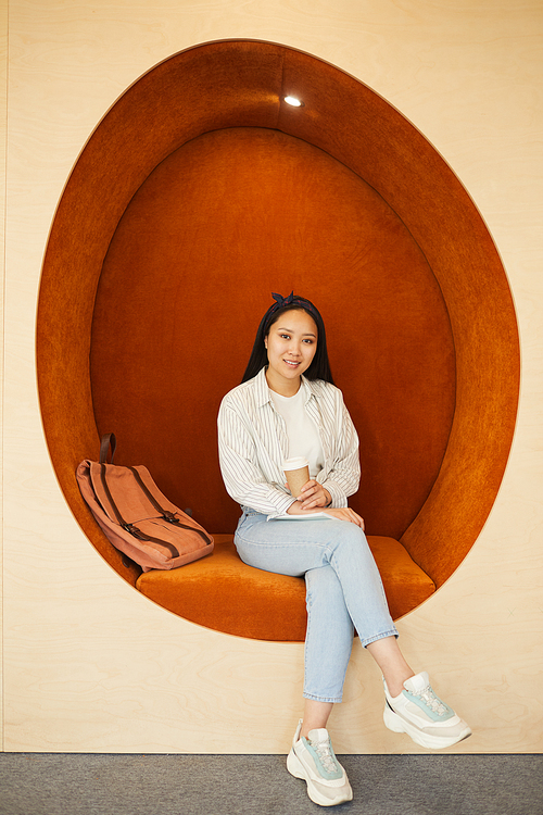 Portrait of smiling pretty Asian student girl in jeans sitting inside of orange armchair and drinking coffee in university hall