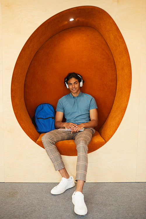 Portrait of happy mixed race student boy in wireless headphones sitting with workbooks in egg-shaped armchair in lobby