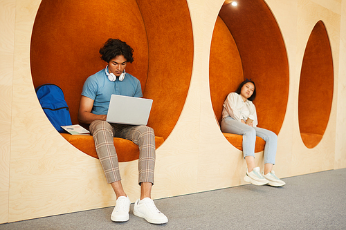 Concentrated mixed race student using modern laptop while preparing task for teacher in campus lobby while his groupmate sleeping on armchair