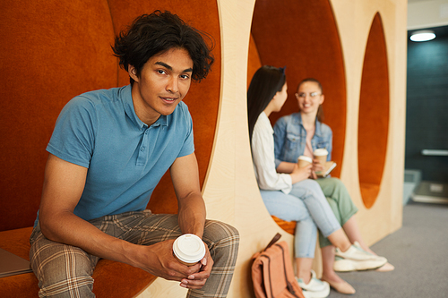 Portrait of smiling handsome dark-skinned boy with disheveled hair sitting in campus lobby and drinking coffee