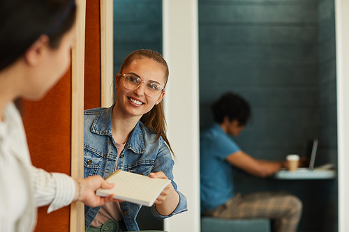 Positive attractive student girl in eyeglasses sharing lecture notes with friend in modern university lobby