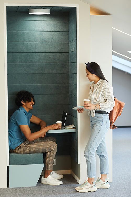 Smiling Asian student girl drinking coffee with mixed race guy while he working on university project in lobby