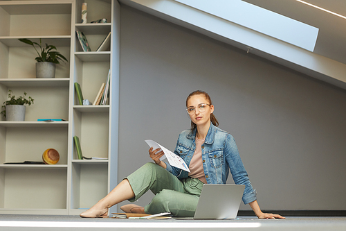 Portrait of serious adult student in eyeglasses sitting on floor at home and learning finance using laptop and papers