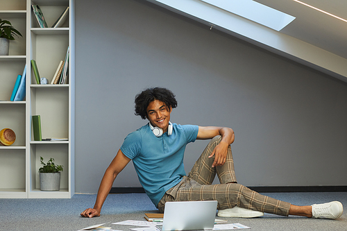 Portrait of smiling stylish student guy with disheveled hair sitting among papers and laptop in own room