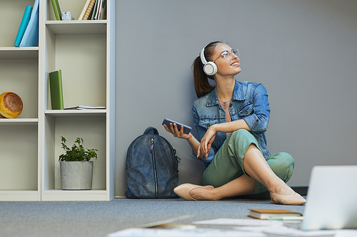 Dreamy attractive student girl sitting on floor and using smartphone while enjoying music in headphones