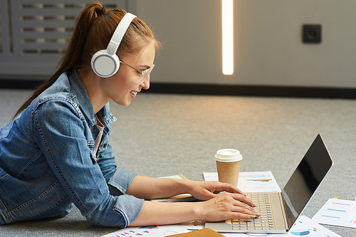 Smiling attractive student girl in wireless headphones lying on floor and using laptop while typing thesis for research project