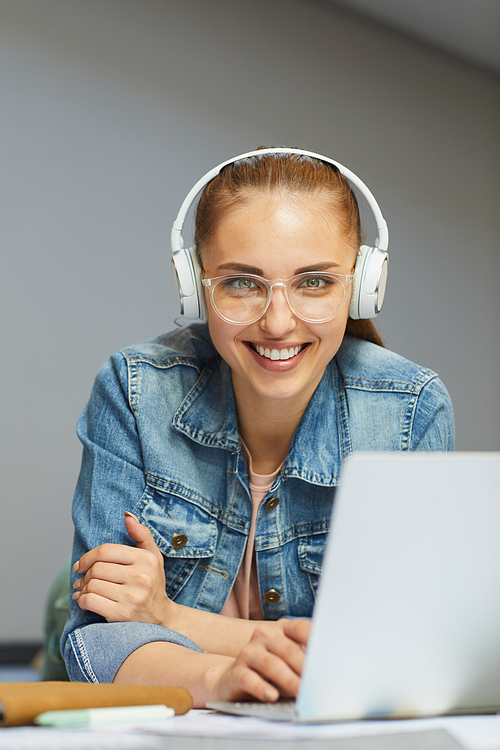 Portrait of cheerful student girl in wireless headphones and eyeglasses using laptop while doing hometask