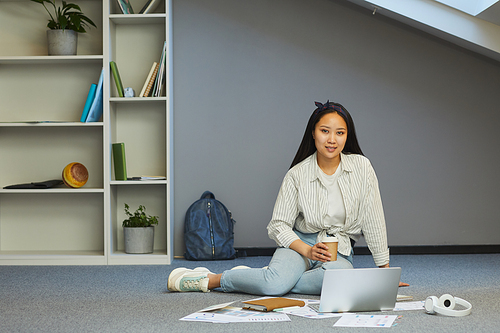 Portrait of smiling Asian girl drinking coffee while working on university task at home