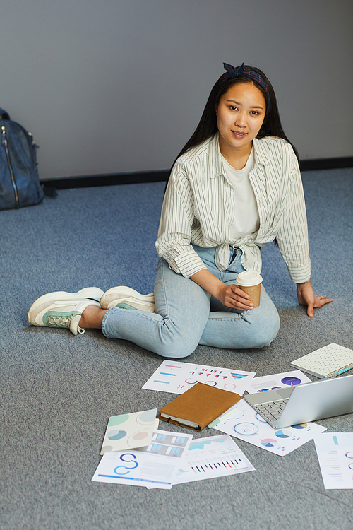 Portrait of smiling Asian girl sitting with coffee on floor and analyzing budget graphs for university coursework