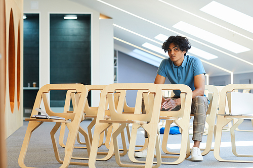 Serious pensive mixed race guy with tousled hair sitting on chair in lecture room while waiting for class