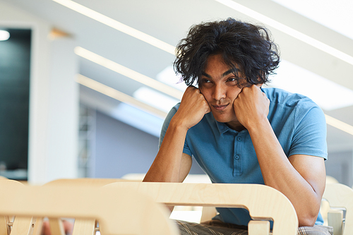 Exhausted mixed race guy in blue tshirt leaning head on fists while listening to boring lecture in classroom