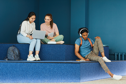 Multi-ethnic university students sitting on steps and using workbooks and devices while doing task before class in blue lobby