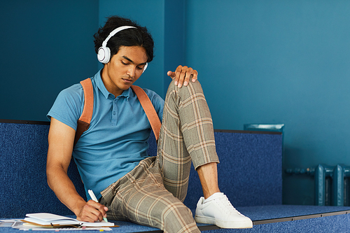 Thoughtful mixed race student boy in checkered trousers checking task in diary and listening to audio in wireless headphones