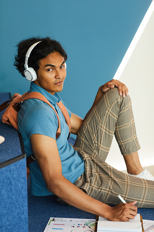 Portrait of content confident mixed race student guy sitting on step in university lobby and making notes in organizer