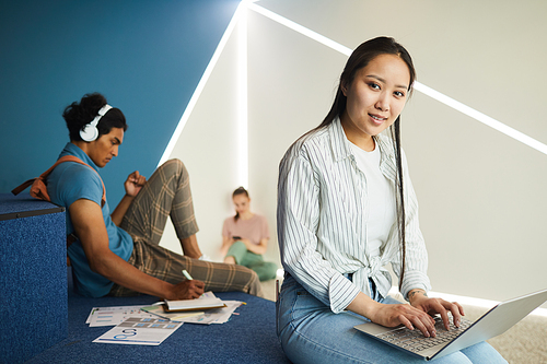 Portrait of smiling Asian student girl in stripped shirt using wifi on laptop while sitting in university hall