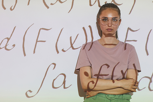 Portrait of serious confident smart girl in eyeglasses standing with crossed arms against projection screen with math formulas