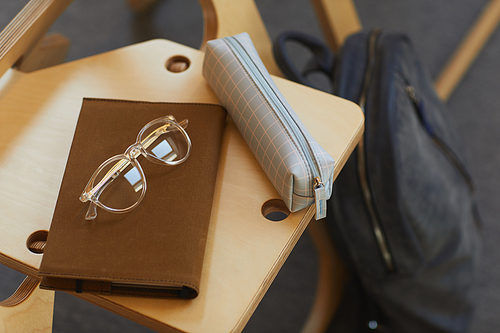 Close-up of checkered pencil case, brown notepad and eyeglasses on wooden chair in university room