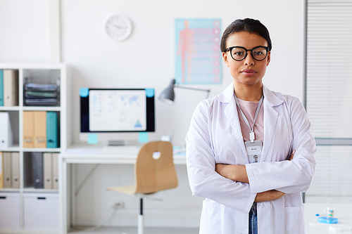 Portrait of young female doctor in eyeglasses standing with her arms crossed and looking at camera at office