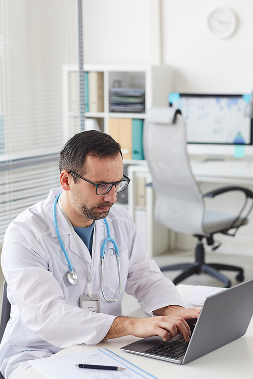 Male mature doctor in white coat sitting at his workplace and working online on laptop at office