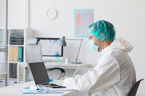 Male doctor in protective clothing sitting at his workplace and typing on laptop computer at office
