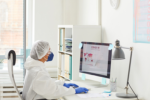 Female doctor in protective clothing sitting at the table in front of the computer monitor and examining the statistics about the coronavirus