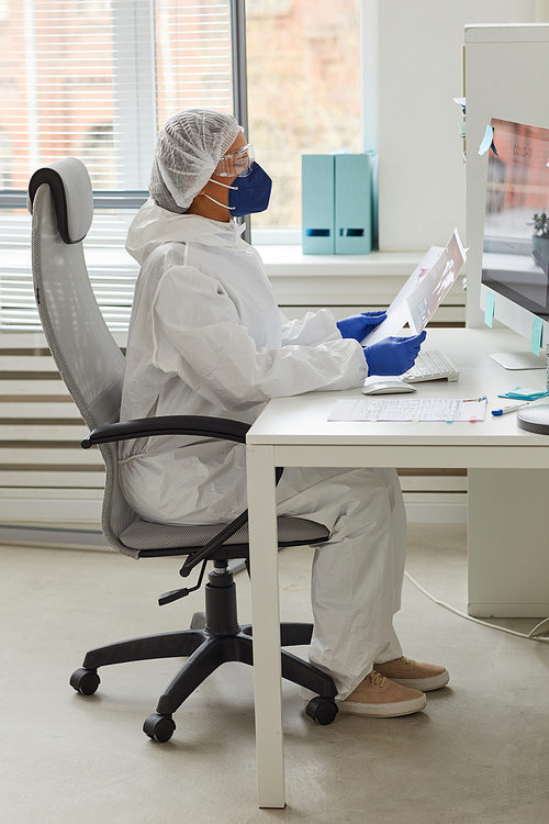 Female doctor in protective workwear sitting at the table working on computer and examining documents at office