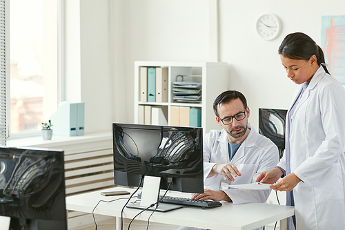 Mature male doctor sitting at his workplace in front of computer and discussing documents with nurse at office