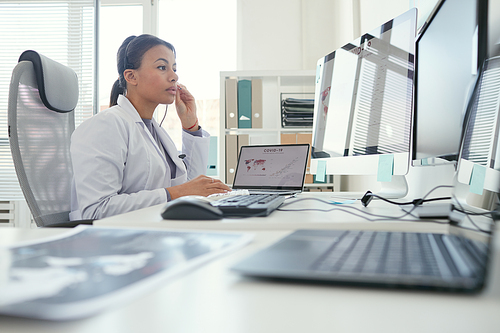 Serious female doctor in headphones looking at computer monitor she has online conference at office