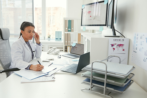 Young female doctor in white coat sitting at the workplace in front of computers and working with documents at office