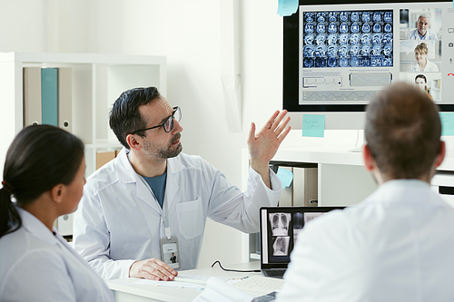 Mature male doctor pointing at x-ray images on computer monitor and discussing them with his colleagues at office