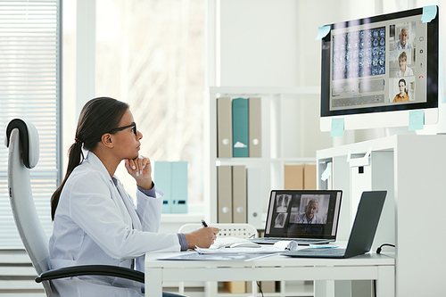Young female doctor sitting at her workplace in front of computer monitor and listening to her colleagues during online conference