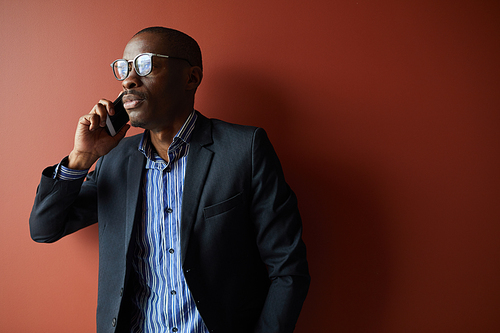 African businessman in eyeglasses and in suit talking on mobile phone while standing against the brown background