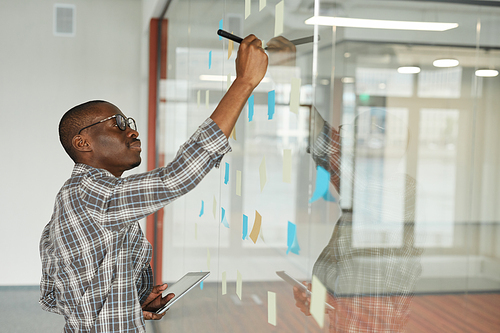 African businessman standing near the glass blackboard and writing plans on adhesive notes at office