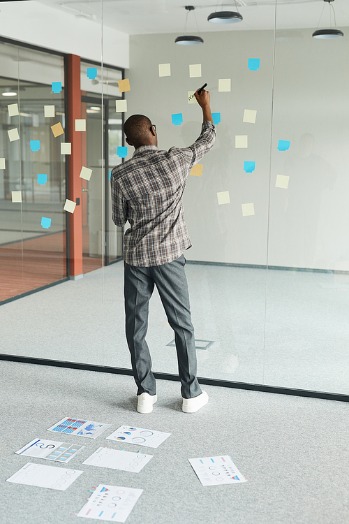 Rear view of African businessman standing in front of glass blackboard and making notes on adhesive notes at office