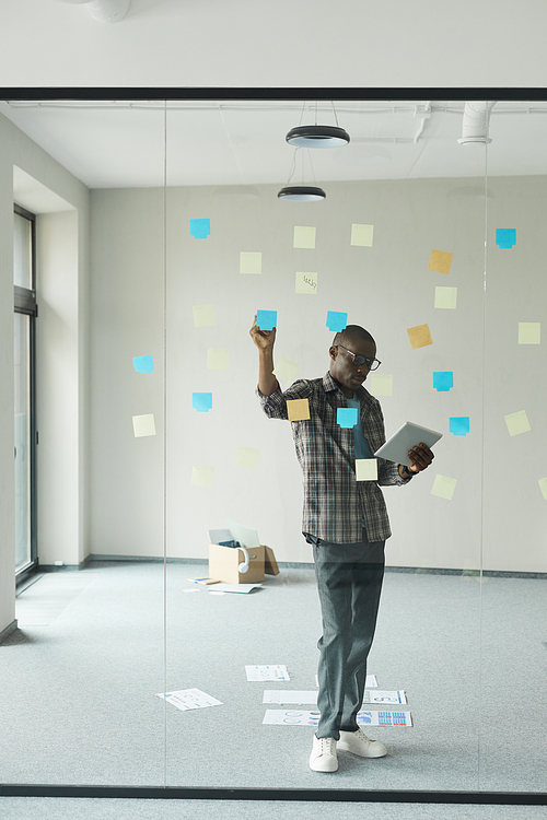 African businessman using digital tablet and making notes on adhesive notes on glass wall at office