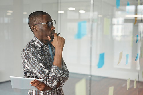 Pensive African businessman in eyeglasses standing in front of glass wall and looking at adhesive notes on it he thinking about new ideas