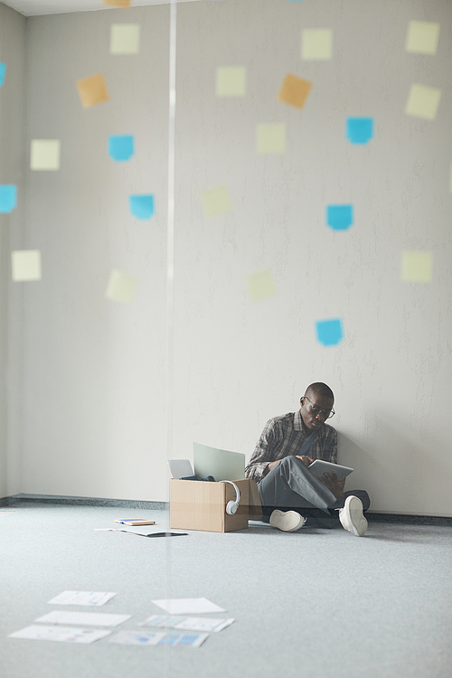 African man sitting on the floor and working online using digital tablet in empty room