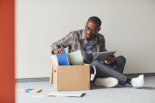 African young man sitting on the floor unpacking the box with documents and using digital tablet