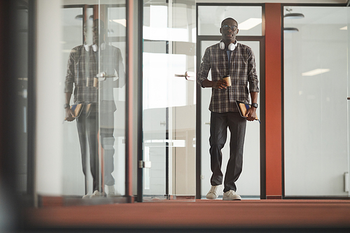 African young businessman in casual clothing walking along the office corridor and drinking coffee