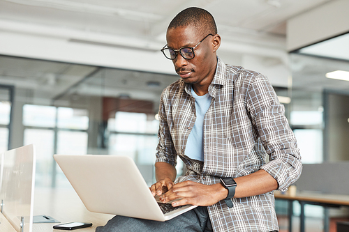 African young designer in eyeglasses typing on laptop computer he working online at office