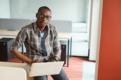 Portrait of African designer in eyeglasses looking at camera while using his laptop computer at work