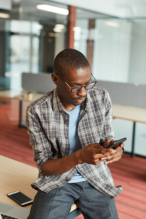 African young man in casual clothing typing a message on his mobile phone while sitting at office