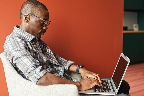 African businessman in casual clothing sitting in armchair and concentrating on his online work he typing on laptop computer