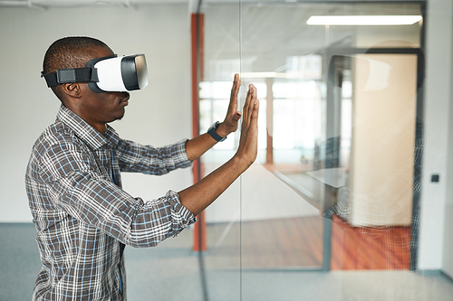 Side view of African man in virtual reality glasses touching the glass wall during virtual reality game in the room