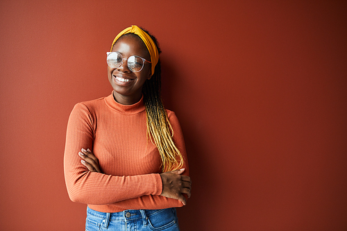 Portrait of African happy woman with stylish hairstyle and in eyeglasses smiling at camera while standing against the brown background