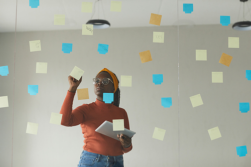 African businesswoman in casual clothing standing in front of the glass wall and writing on adhesive notes the new plans