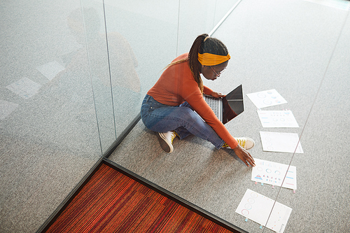 Young designer sitting on the floor and examining documents with charts using her digital tablet at office