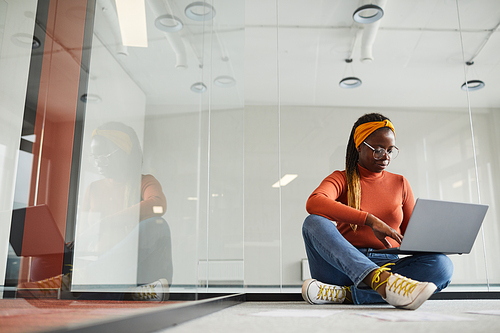 Designer in casual clothing sitting on the floor and working online using her laptop computer at modern office