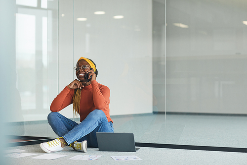 African female designer sitting on the floor with laptop and talking on mobile phone in empty office